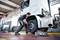 A mechanic is changing a tyre on a lorry in a workshop. The lorry is jacked up, and the mechanic is wearing work gloves.