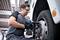 A man is changing a vehicle's tyre using a cordless impact driver in a workshop, wearing safety glasses and gloves.