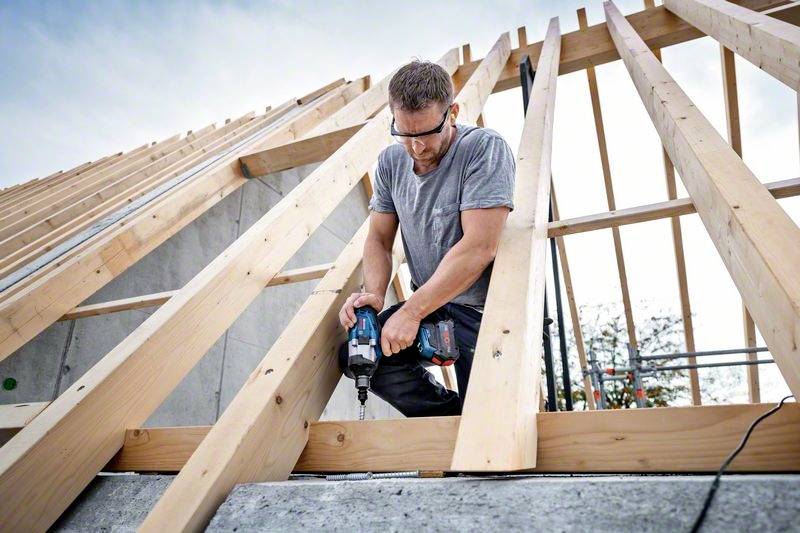 A man is using a cordless drill driver to secure wooden battens. He is working on a roof truss frame outdoors.