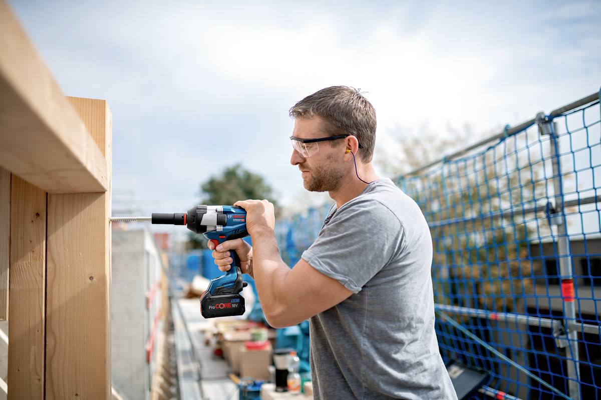 A man is working with an electric drill on a construction site. He is wearing safety glasses and ear plugs.