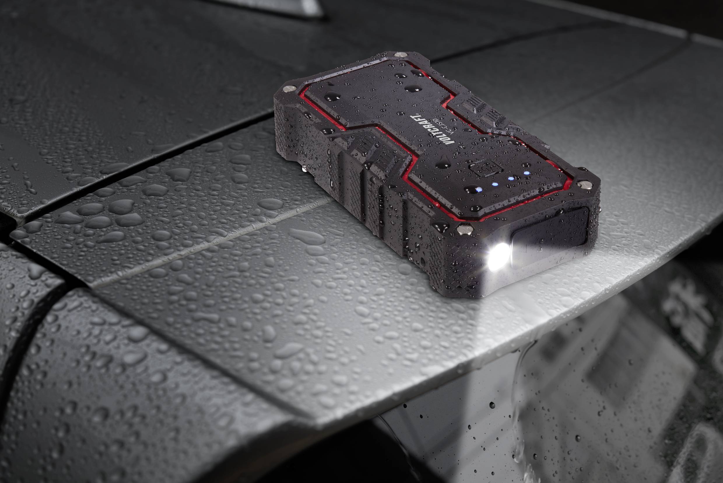 A portable charger with an LED light sits on a wet car roof. It has a robust design with a black and red casing.