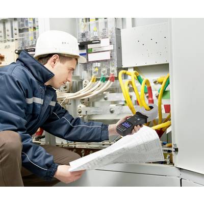 A technician wearing a hard hat is checking electrical connections in a control cabinet. He is holding a measuring device and construction plans in his hand.
