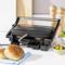 A contact grill on a kitchen worktop is grilling burger patties. In the background are salt and pepper shakers and a bowl of pickled gherkins.