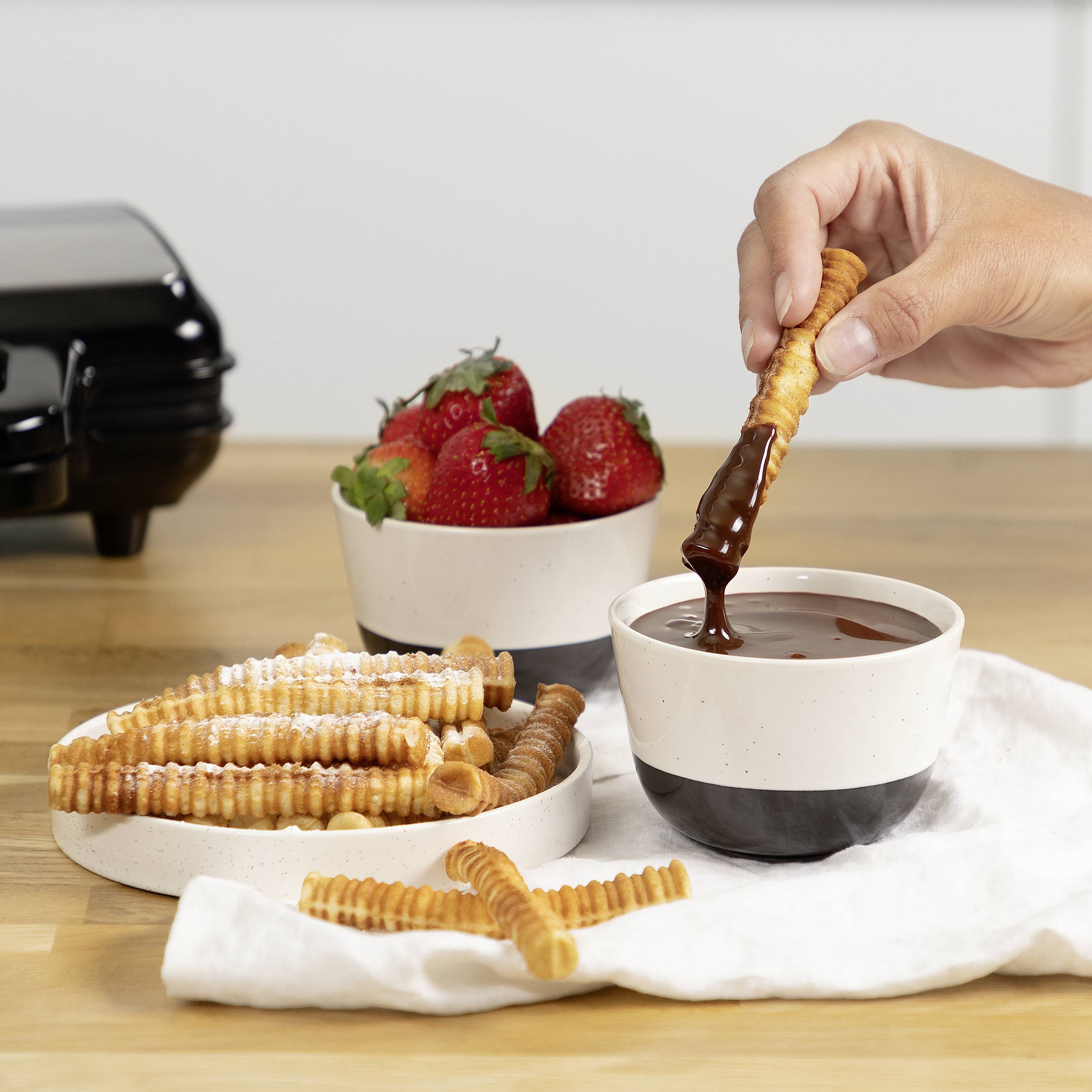 A hand dips a cracker into liquid chocolate. In the background, there is a bowl of strawberries and a toaster.