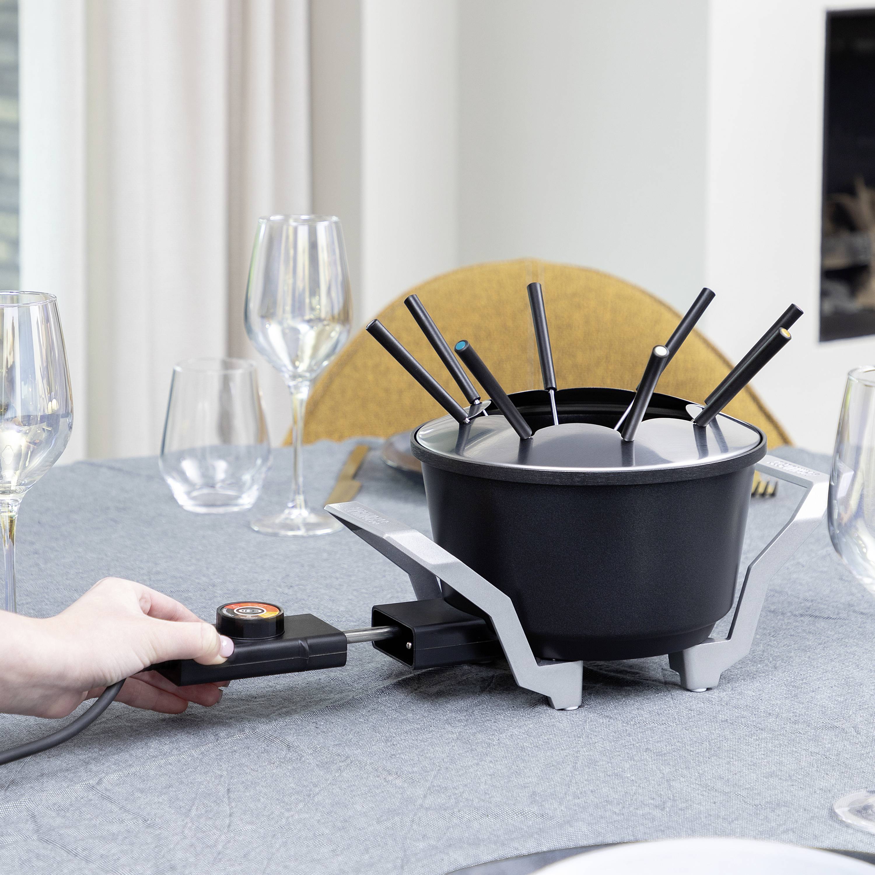 A person is preparing an electric fondue pot on a laid table, surrounded by wine glasses and plates.