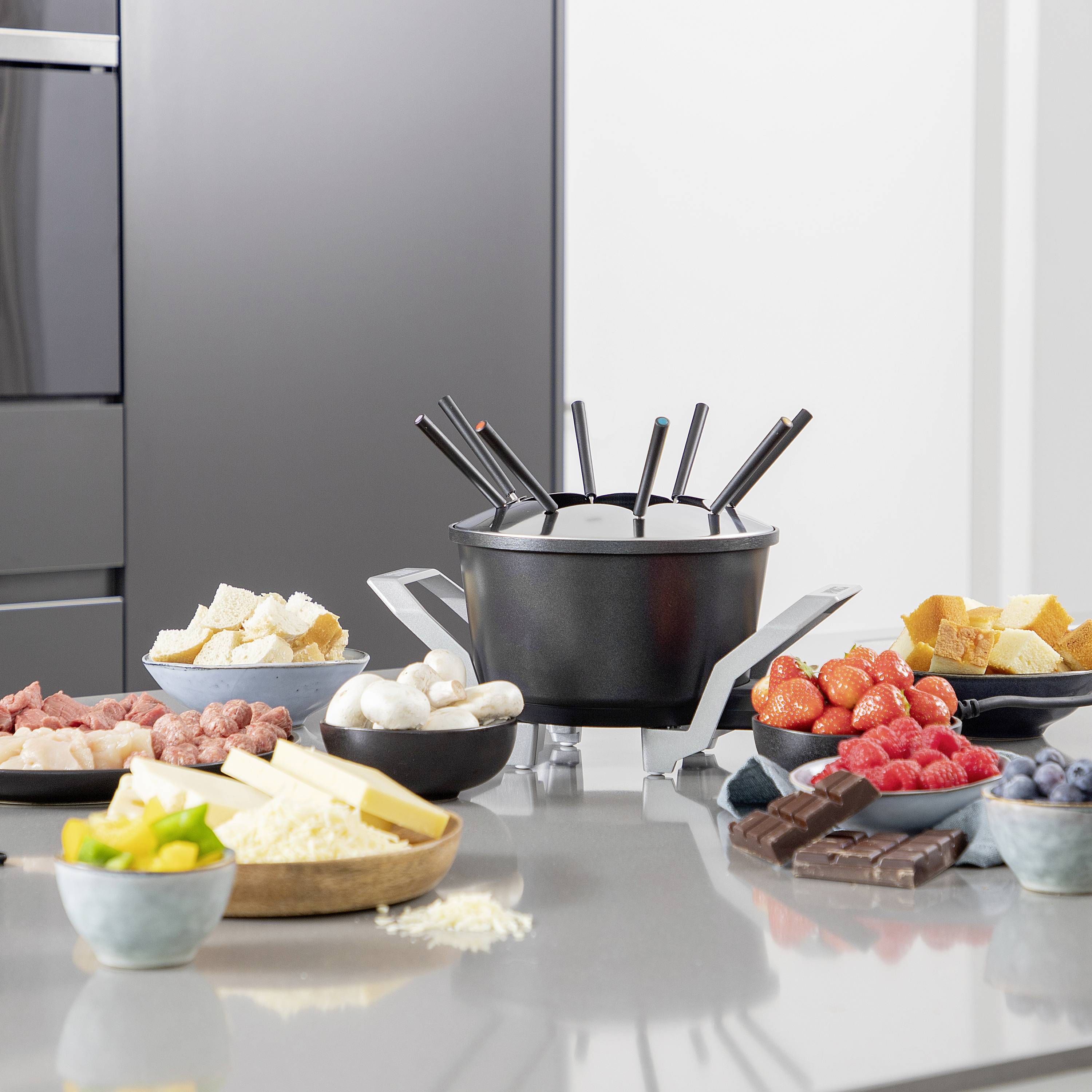 Fondue table with a heated pot, surrounded by meat, vegetables, bread, strawberries, blueberries, chocolate and cheese for a meal.