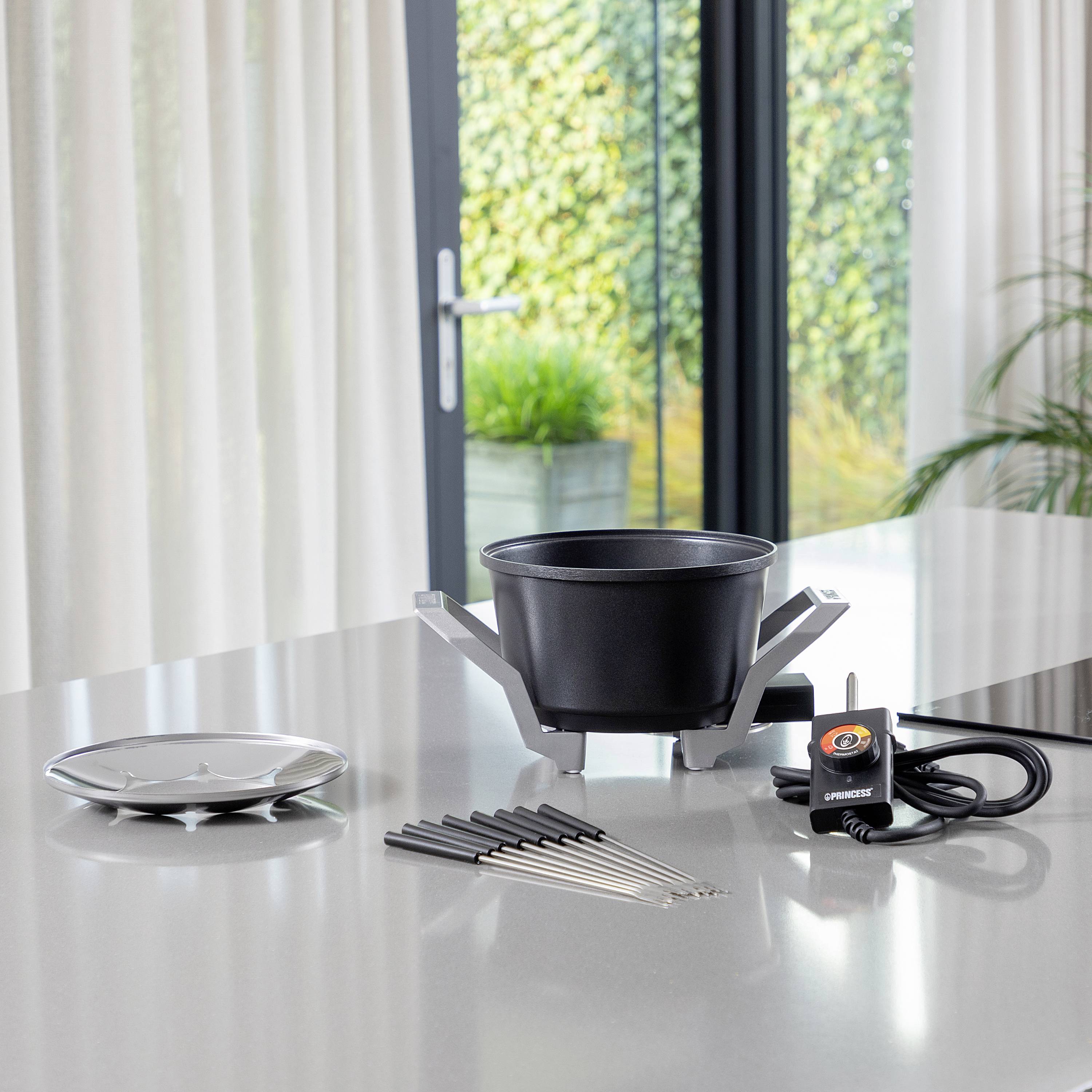 A fondue pot set on a table with chopsticks, burner, and trivet. A glass door with plants visible outside in the background.
