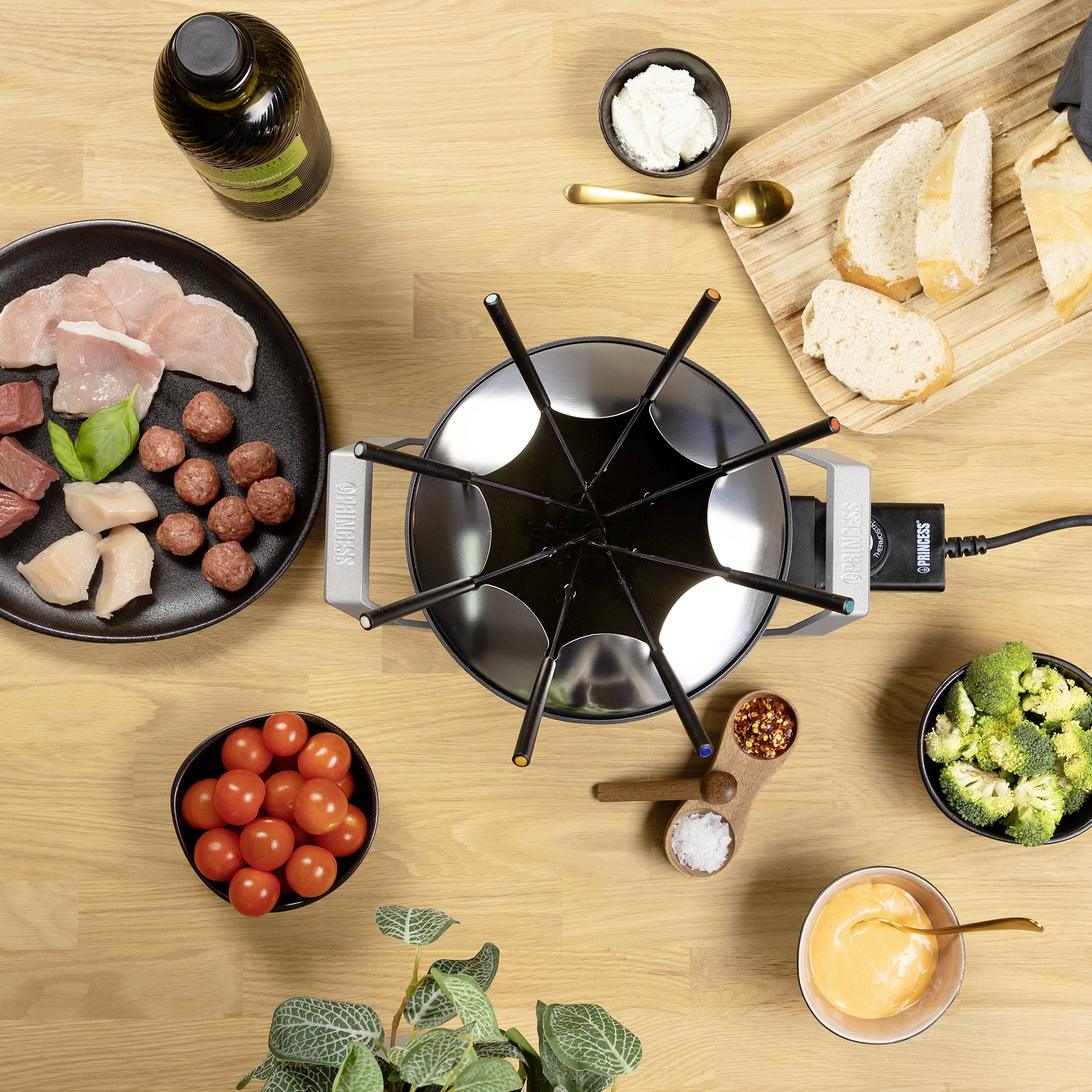 A fondue scene with sliced meat, bread, tomatoes, broccoli and dips. A fondue pot sits in the centre on a wooden table.