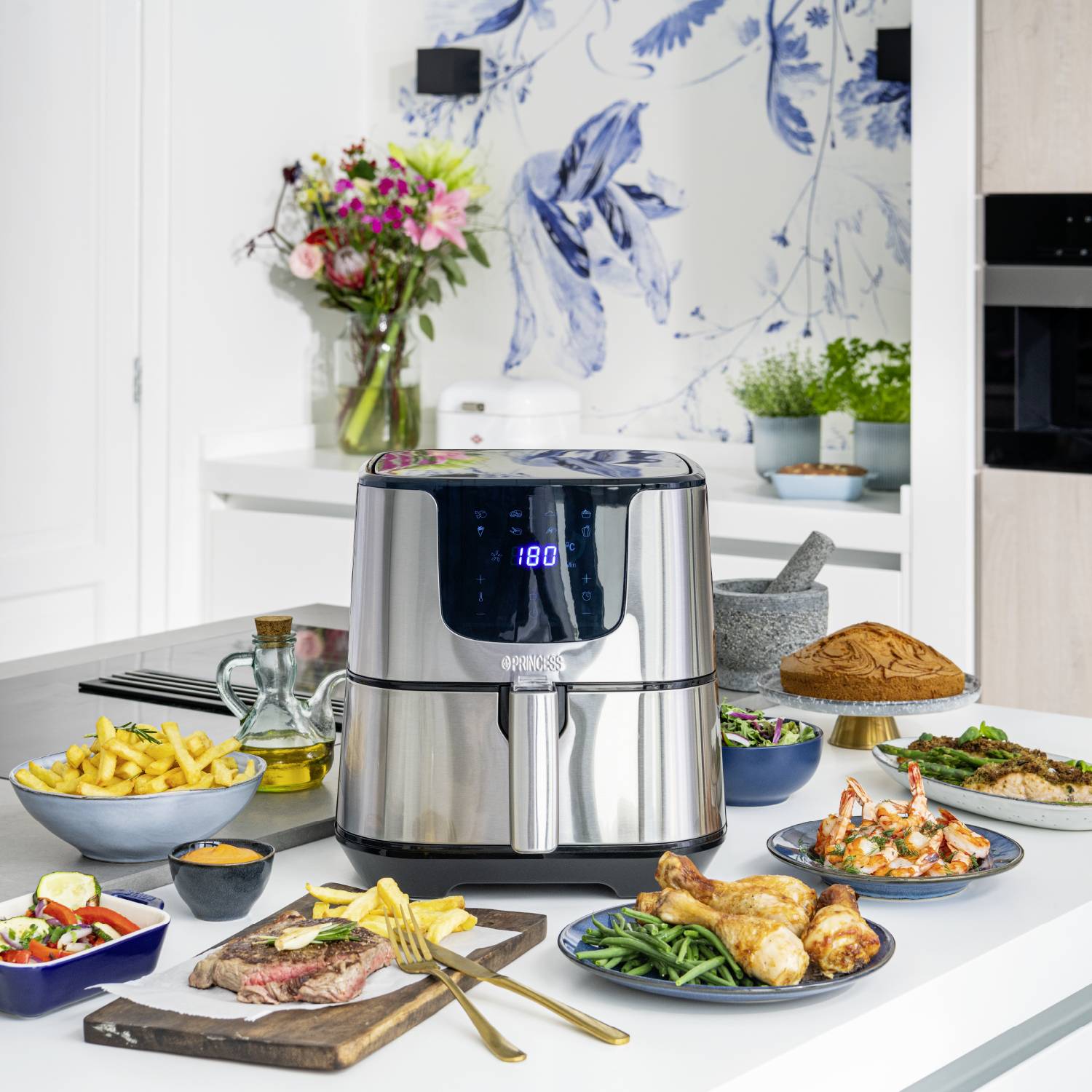A modern air fryer on a kitchen table, surrounded by various dishes such as chicken, chips, and vegetables. Flowers in the background.