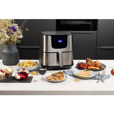 A hot air fryer is sitting on a kitchen worktop, surrounded by chips, cake, chicken legs, and fish dishes.