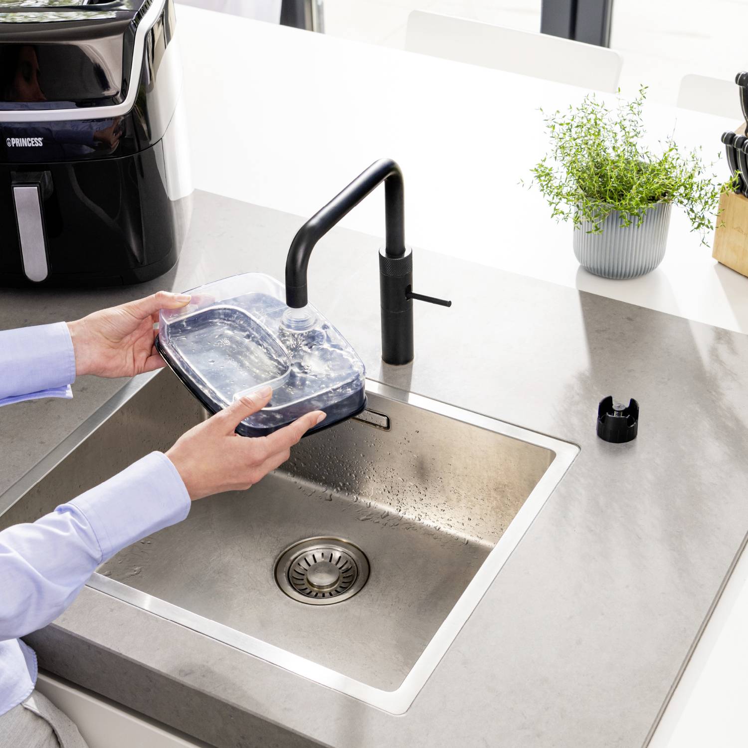 'A person fills a water container at the sink. An air purifier sits on the worktop. A plant stands nearby.'