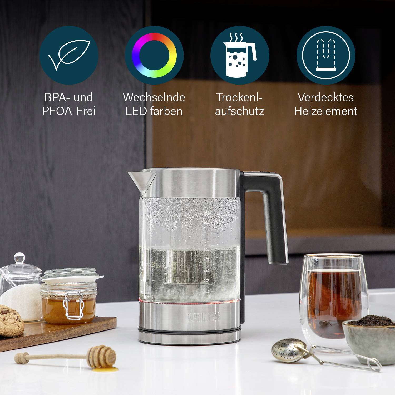 A glass and stainless steel kettle in a kitchen, surrounded by honey, tea, and biscuits. Caption: 'Changing LED colours, BPA and PFOA free, Dry-run protection, Concealed heating element'.
