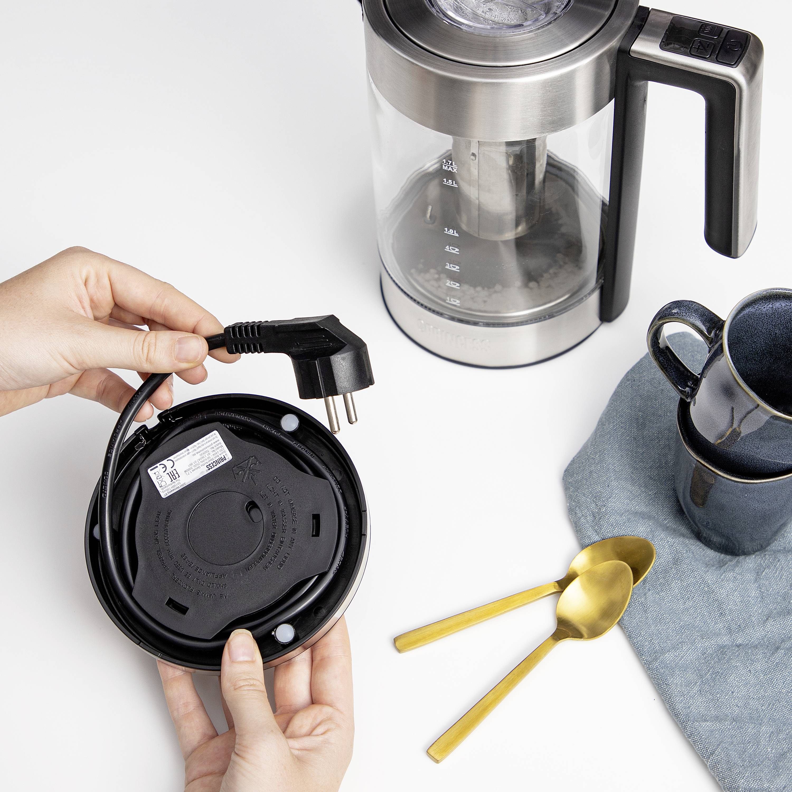 A person is holding the plug of an electric kettle next to a jug. Beside it are a blue mug and a golden spoon.