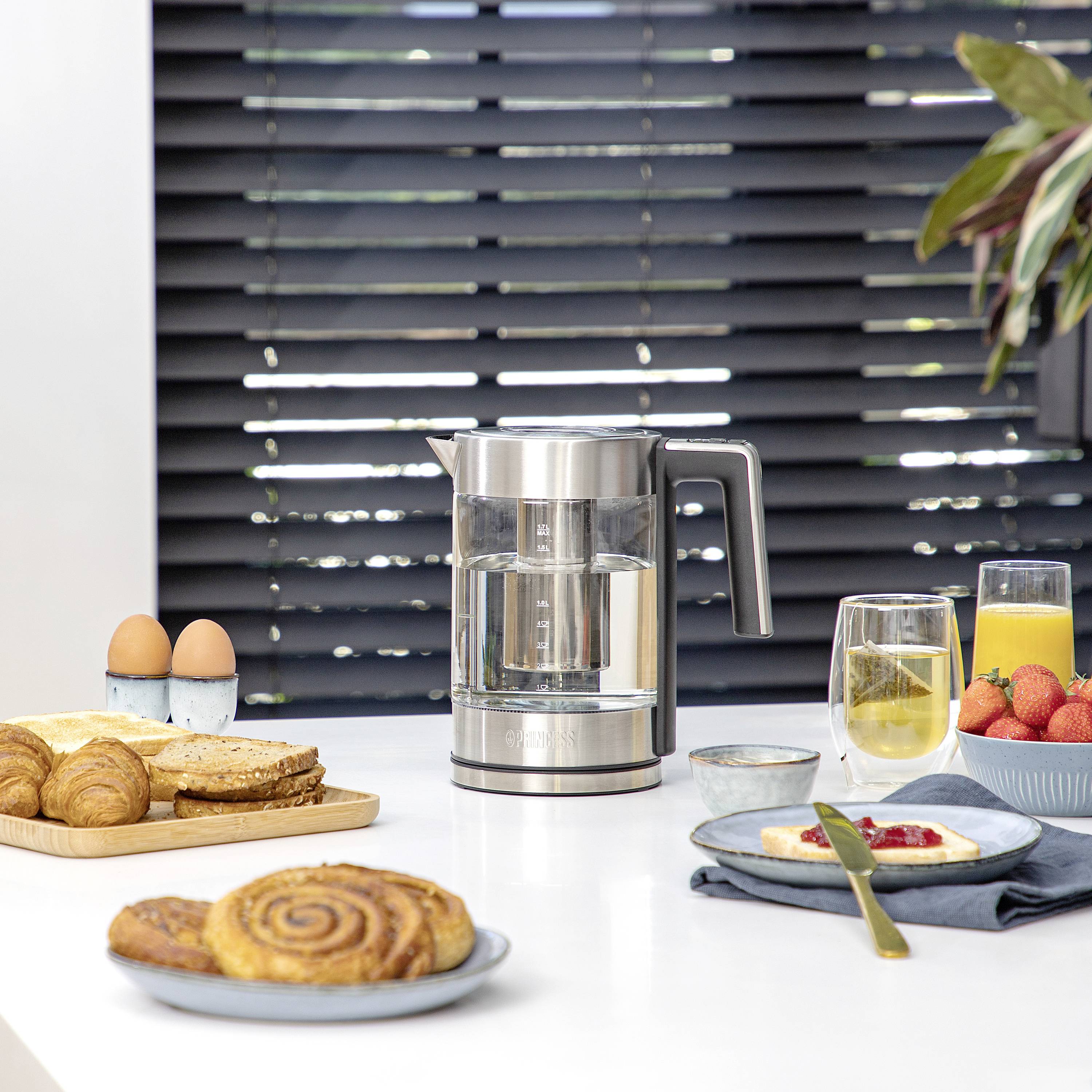 Electric kettle beside a breakfast table with croissants, bread rolls, orange juice and strawberries on a modern, bright kitchen table.