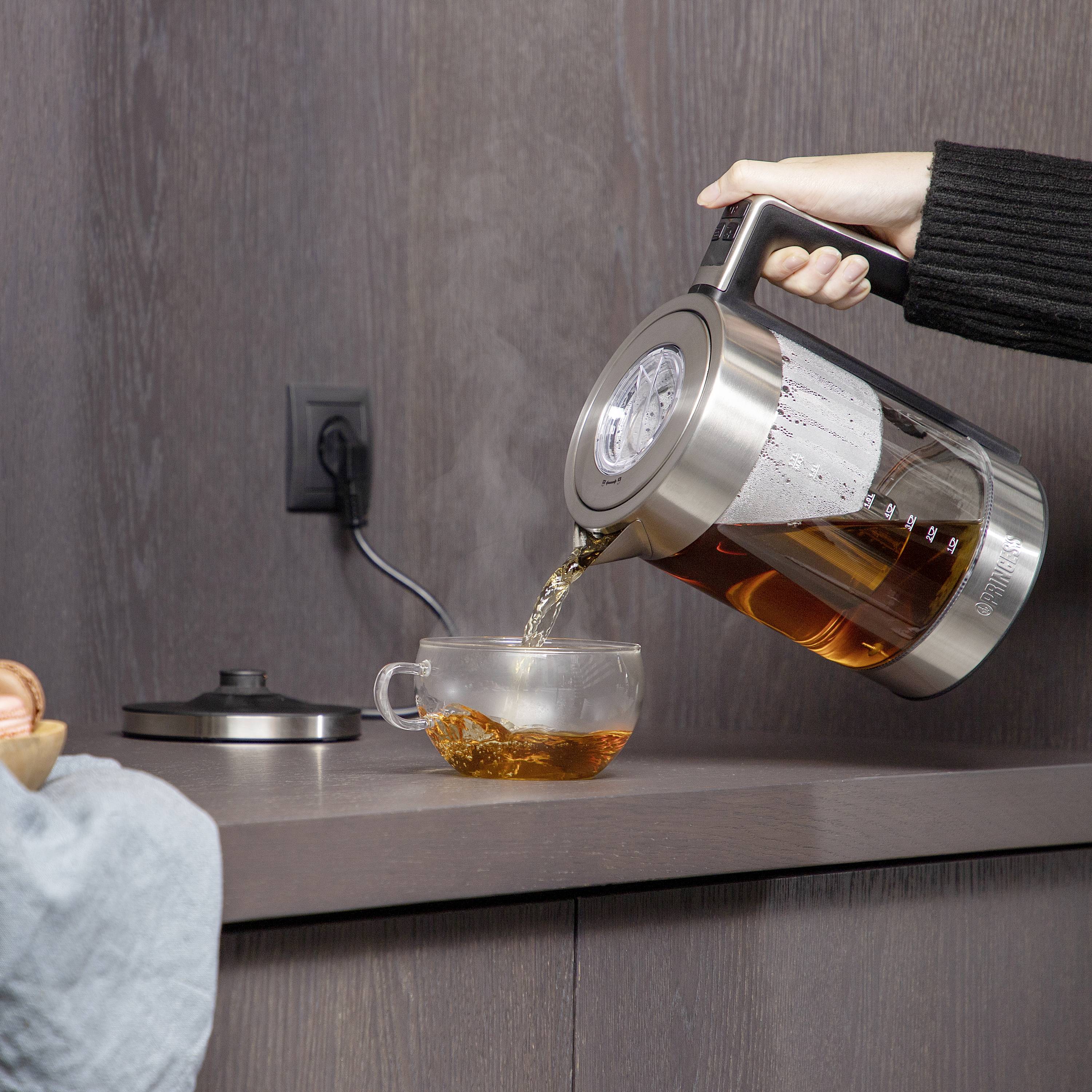 A person is pouring hot tea from a kettle into a mug on a kitchen worktop. A power socket is visible in the background.