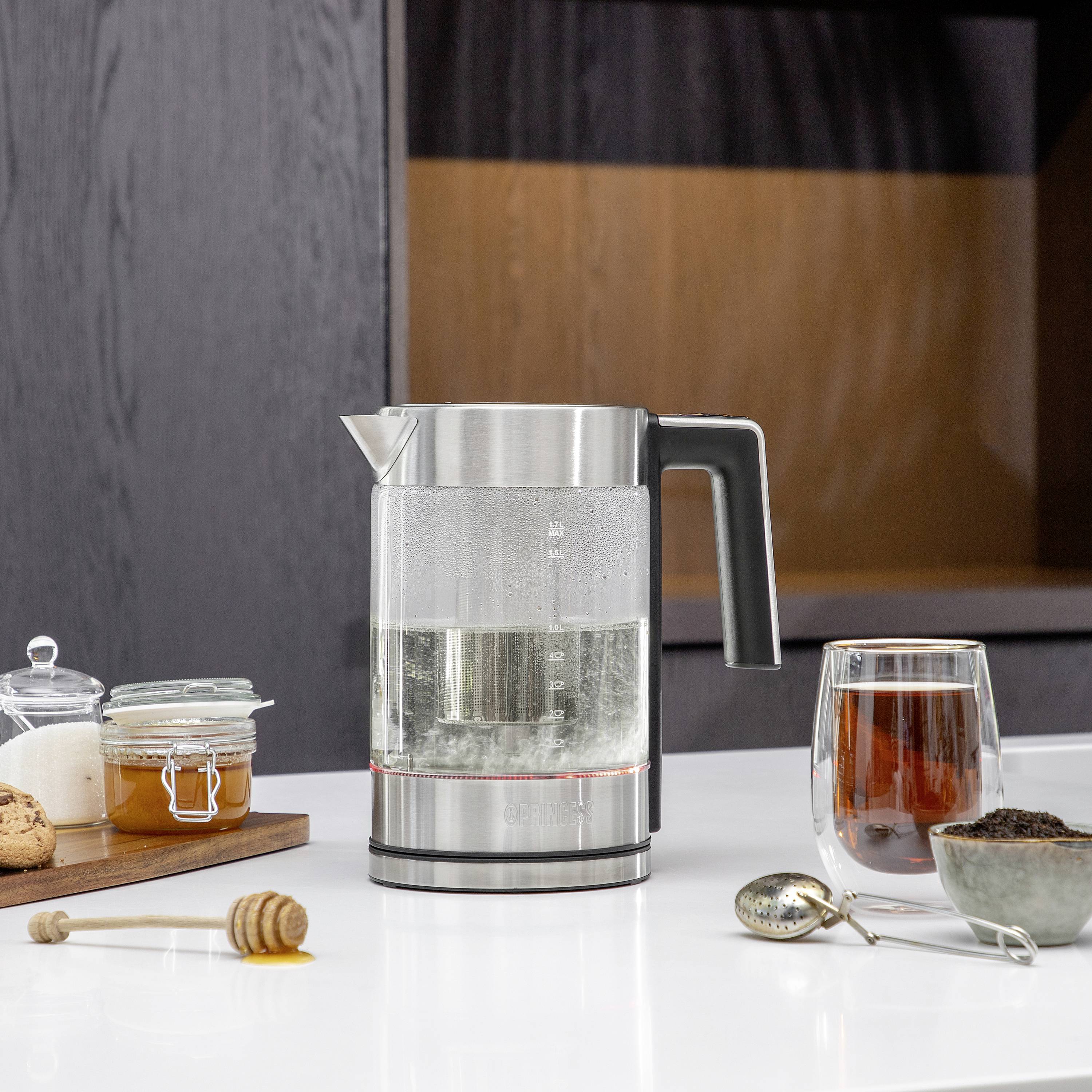 A kettle with water on a table next to a cup of tea, a spoon with tea leaves, honey and a biscuit.