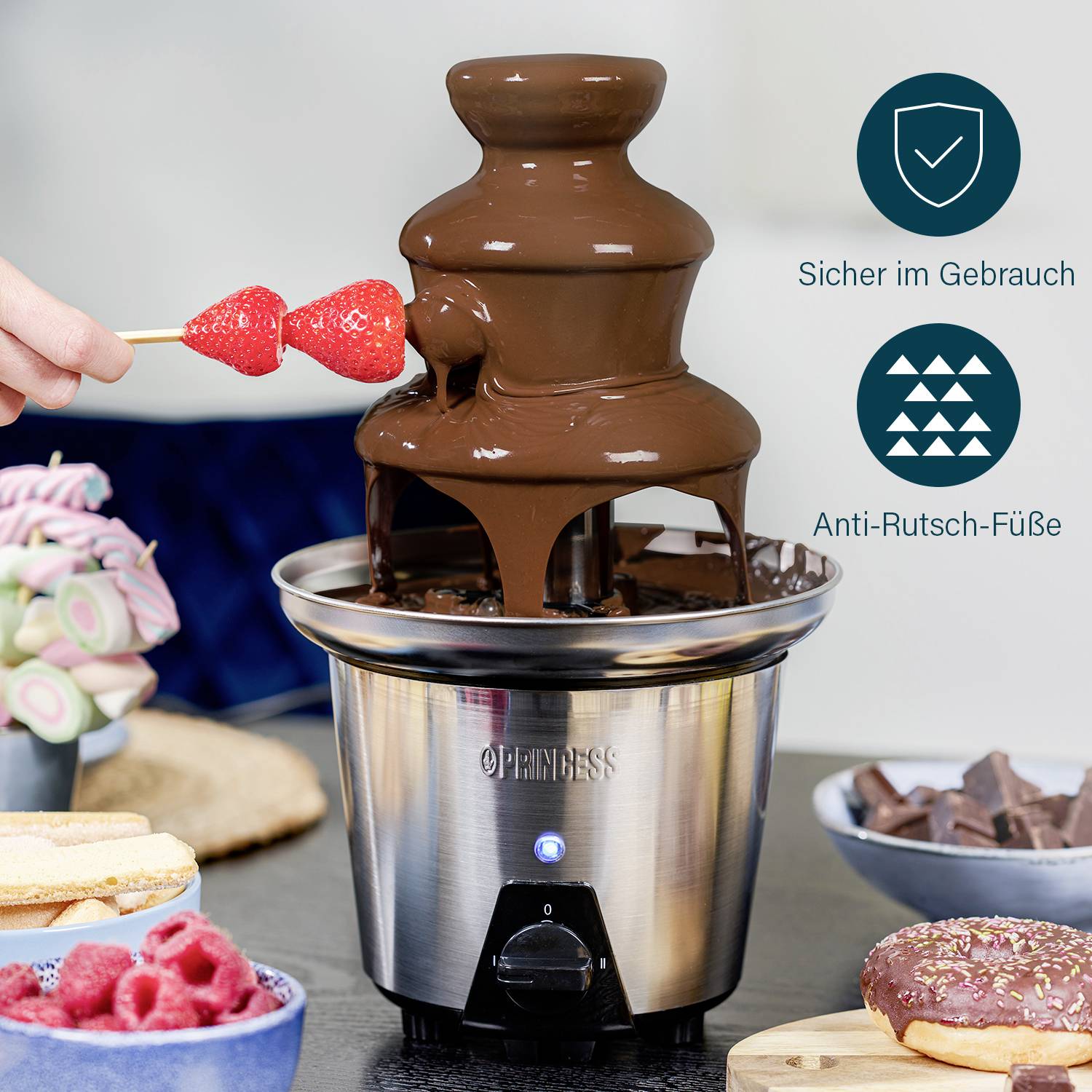 Chocolate fountain with liquid chocolate, a hand holding strawberries. Bowls of sweets in the background.