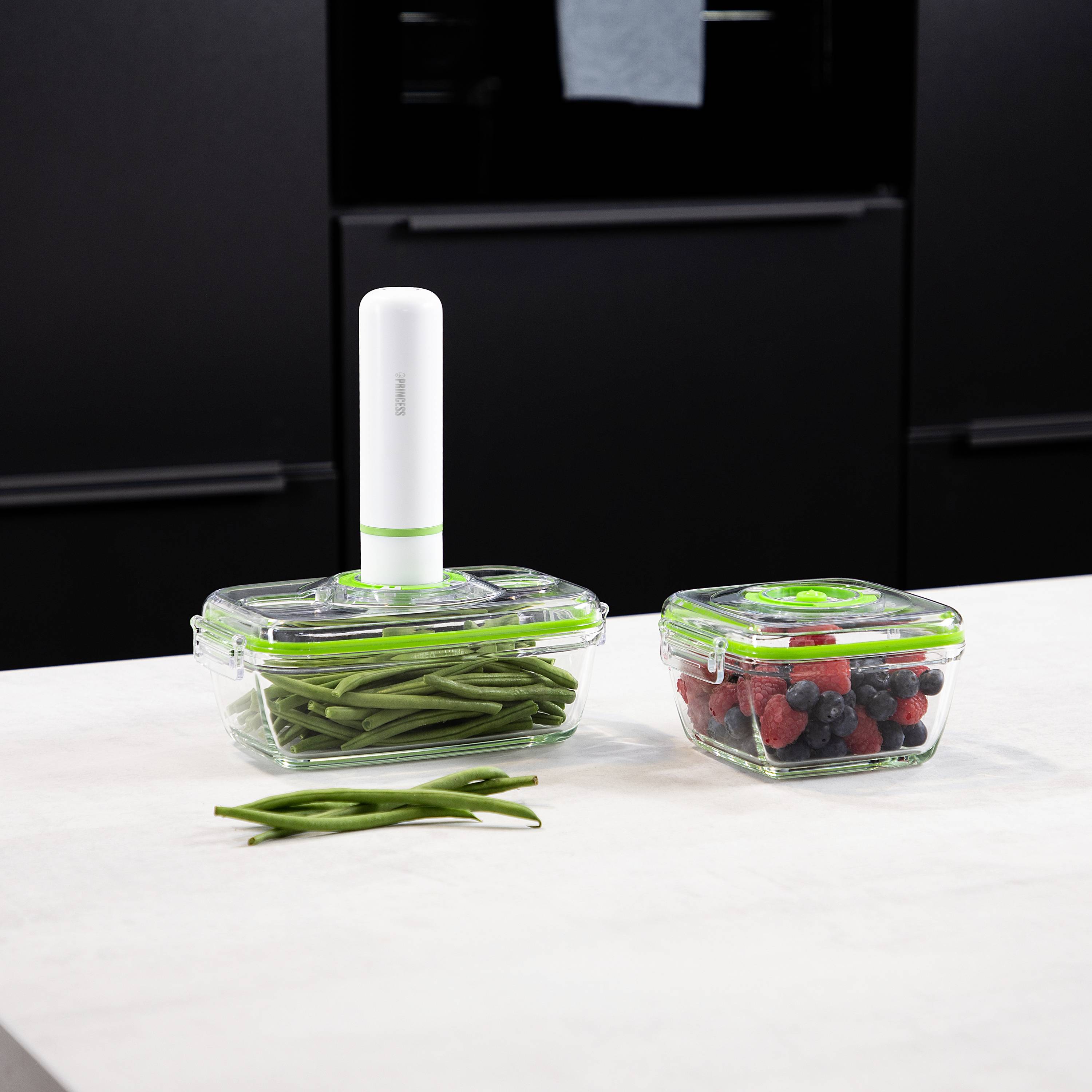 Two food storage containers on a table, one with green beans and one with mixed berries, with a vacuum sealing system underneath.