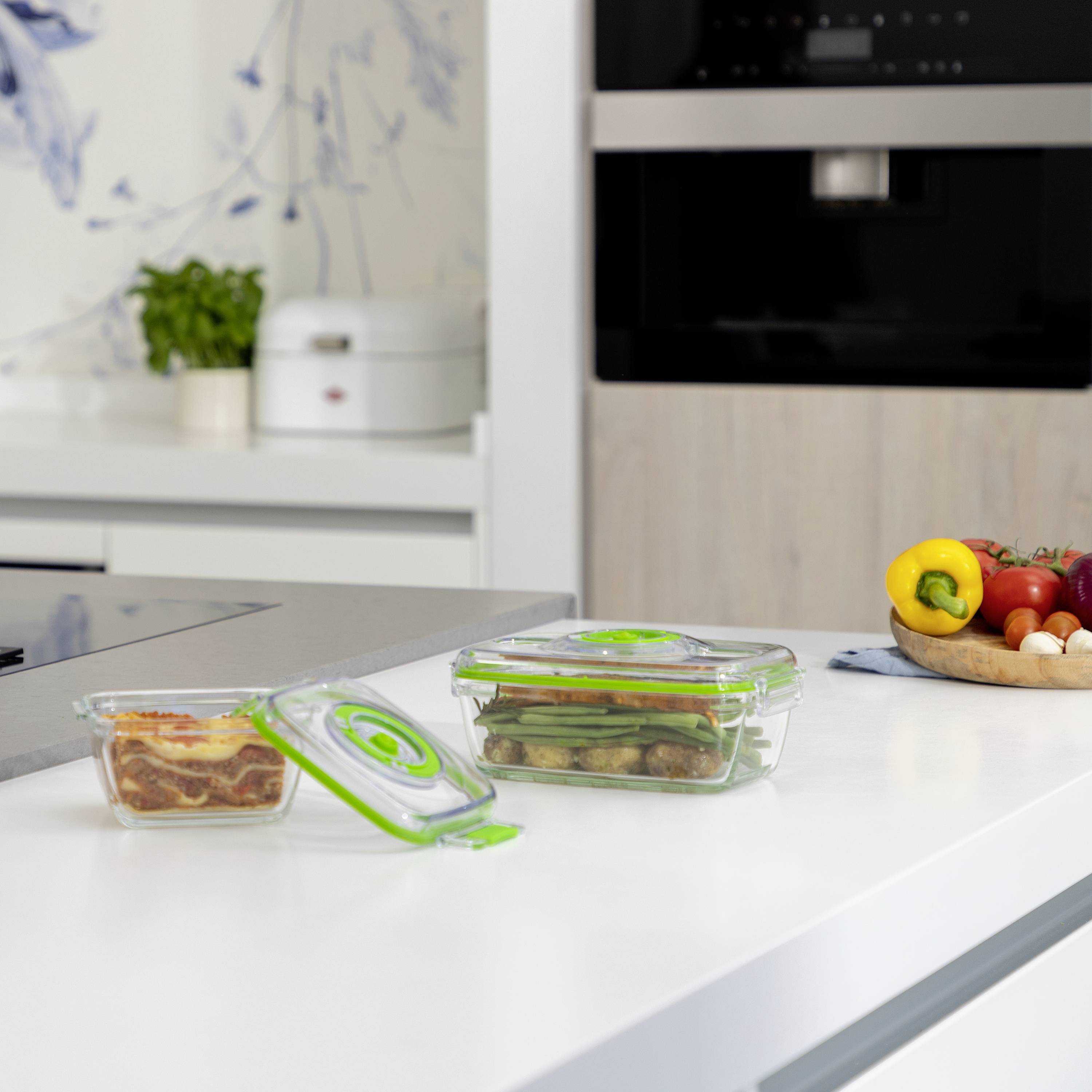 Food boxes with vegetables and lasagne on a light kitchen countertop. A coffee machine and a vegetable basket are visible in the background.