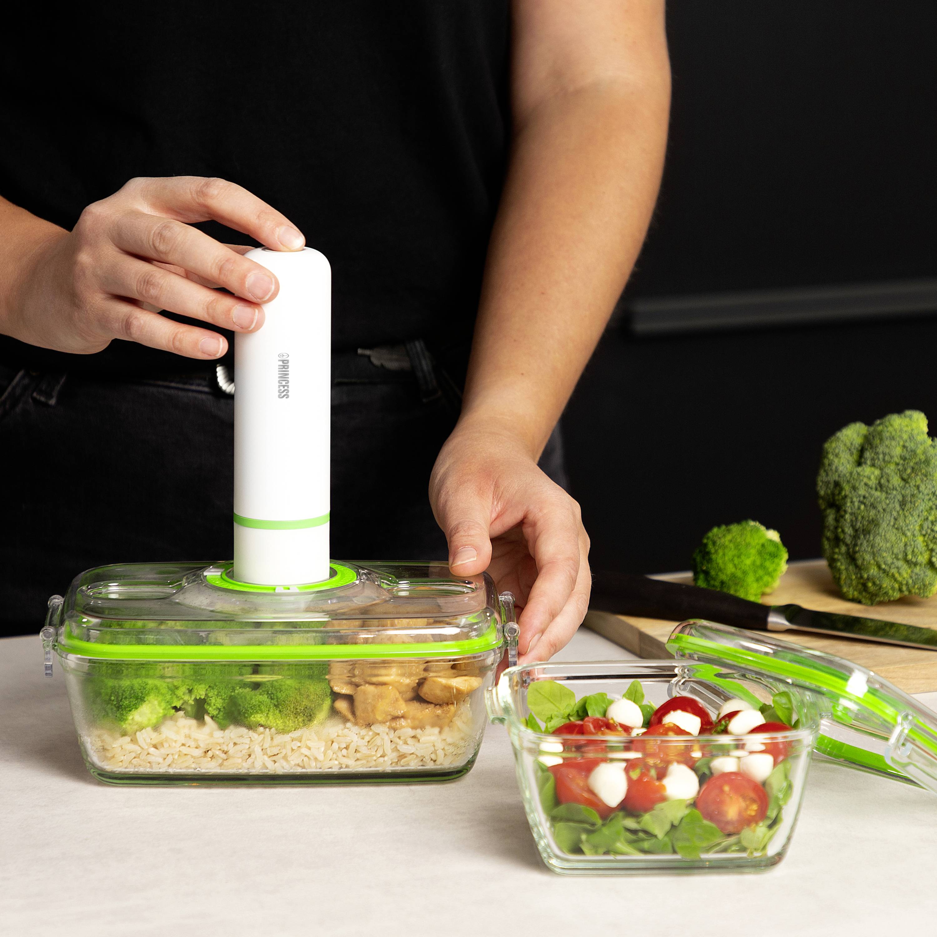A person is using a vacuum sealer to seal a food storage container with broccoli and rice. A salad in a glass bowl is placed beside it.