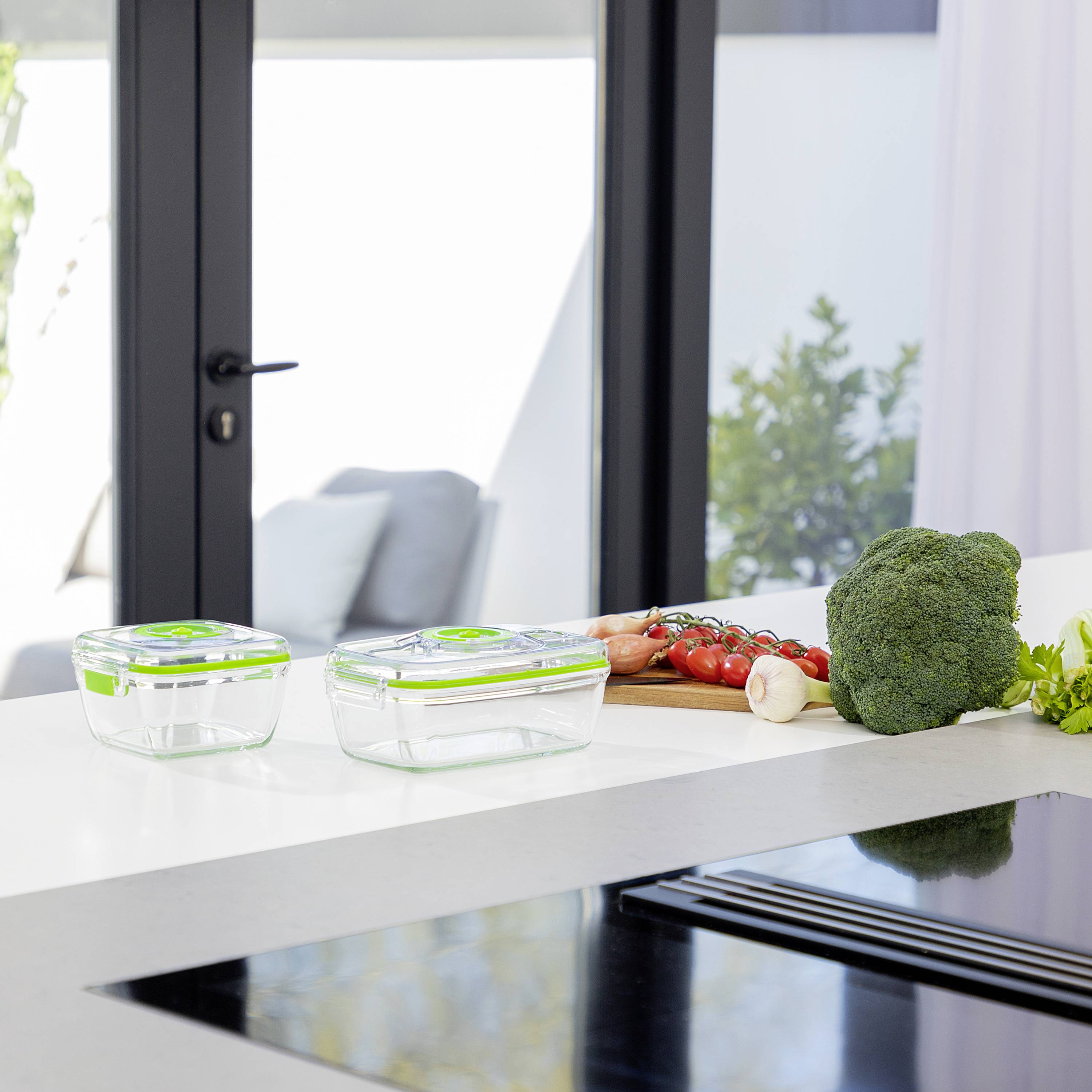A modern kitchen with a white worktop. Two glass containers on the left, vegetables on the right: tomatoes, broccoli. Bright windows in the background.