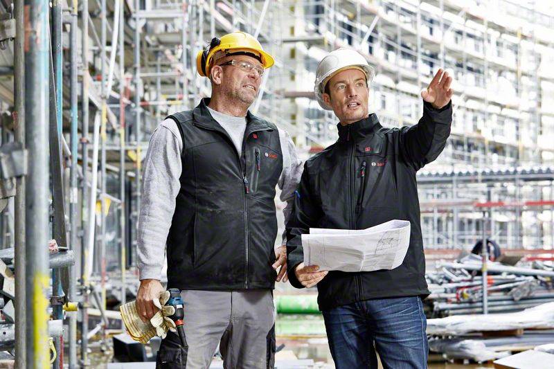 Two construction workers wearing hard hats stand on a building site. One is holding building plans, and both are looking attentively into the distance. Scaffolding is visible in the background.