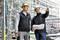 Two construction workers wearing hard hats stand on a building site. One is holding building plans, and both are looking attentively into the distance. Scaffolding is visible in the background.