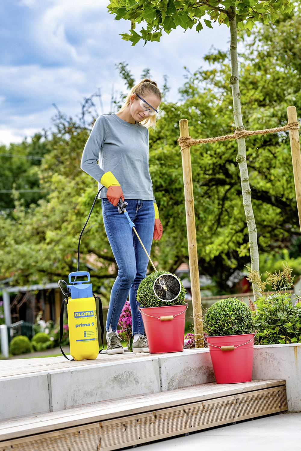 A person is watering plants in red pots on a terrace using a spray device. Trees can be seen in the background.