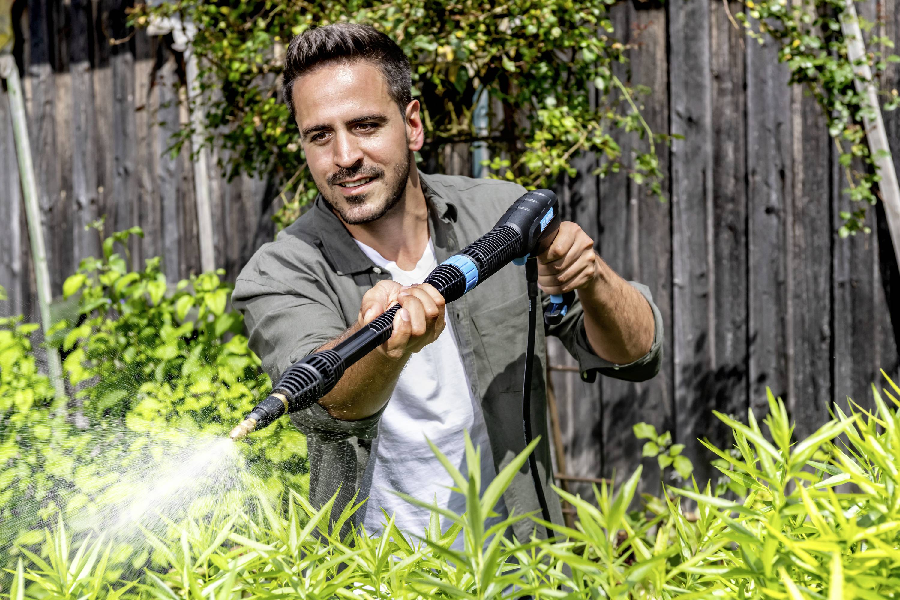 A man is spraying water on plants in the garden, surrounded by green foliage and a wooden fence in the background.