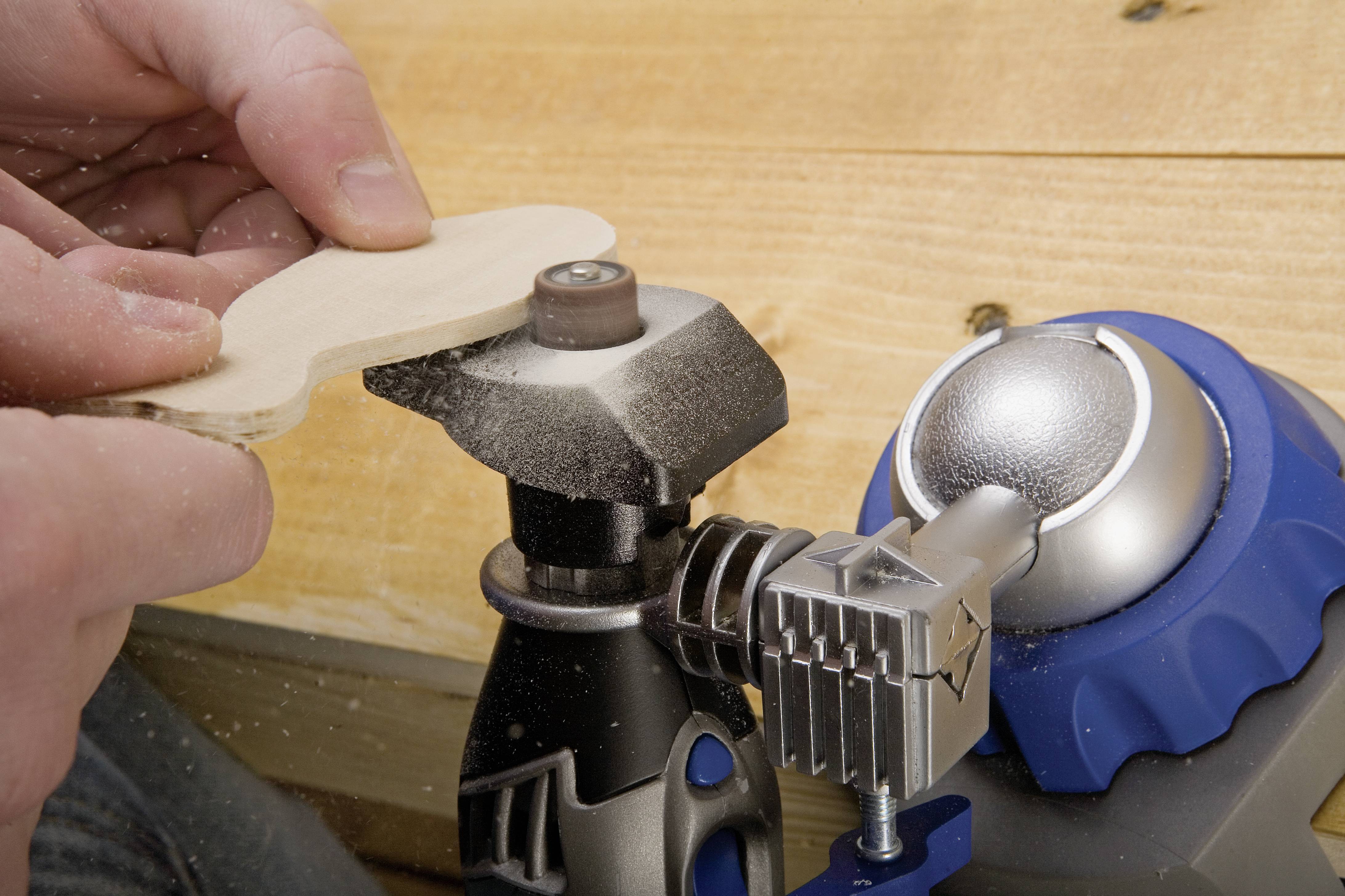 A person is sanding a piece of wood with an electric sanding tool. Wood shavings are visible as the wood is being worked on.