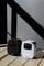 Two compact speakers, one black and one white, stand on a wooden floor in sunlight, casting diagonal shadows.