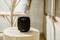 Small black speaker with a display on a wooden table, showing a music track. A rattan chair is in the background.