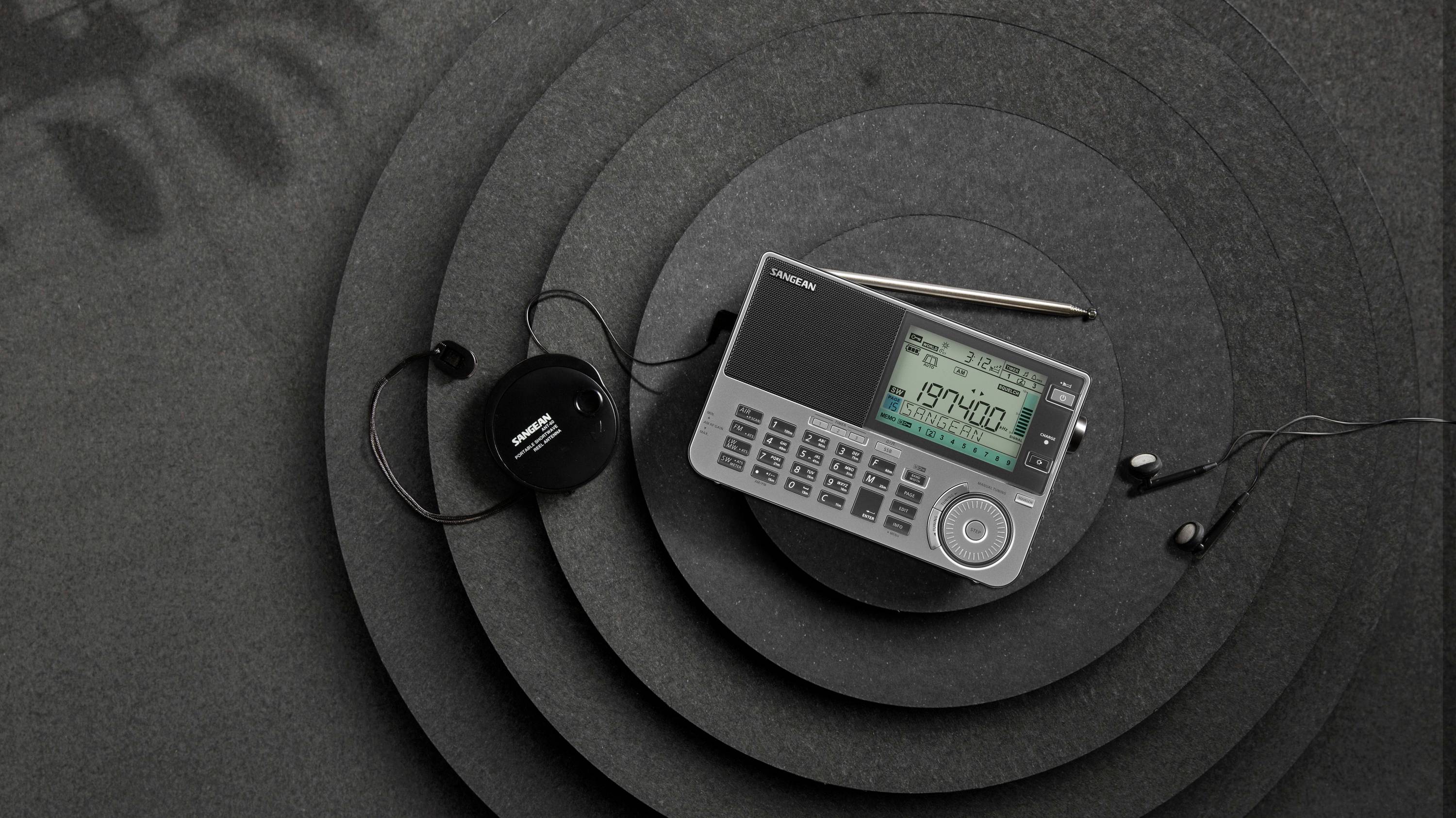 A modern, portable radio with a display and keypad sits on concentric black circles, alongside headphones and an antenna.