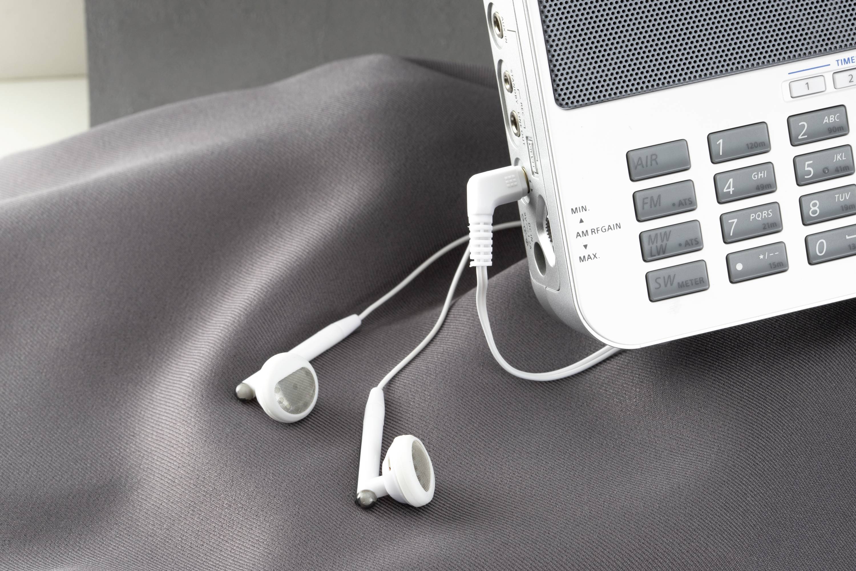 A radio with connected white earphones lies on grey fabric. The focus is on the earphones and the control panel of the radio.