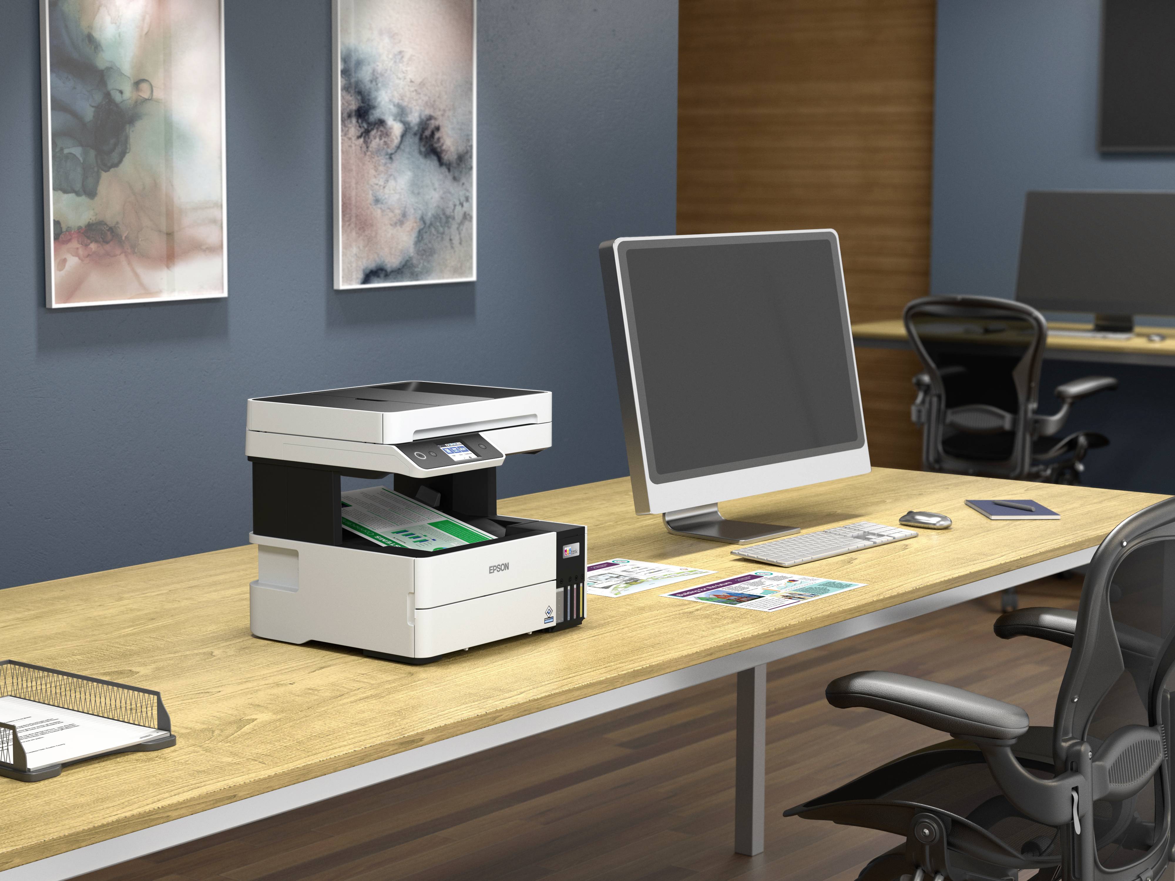 A printer sits on a wooden desk, surrounded by a computer, chairs and paper documents in a modern office.
