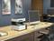 A printer sits on a wooden desk, surrounded by a computer, chairs and paper documents in a modern office.