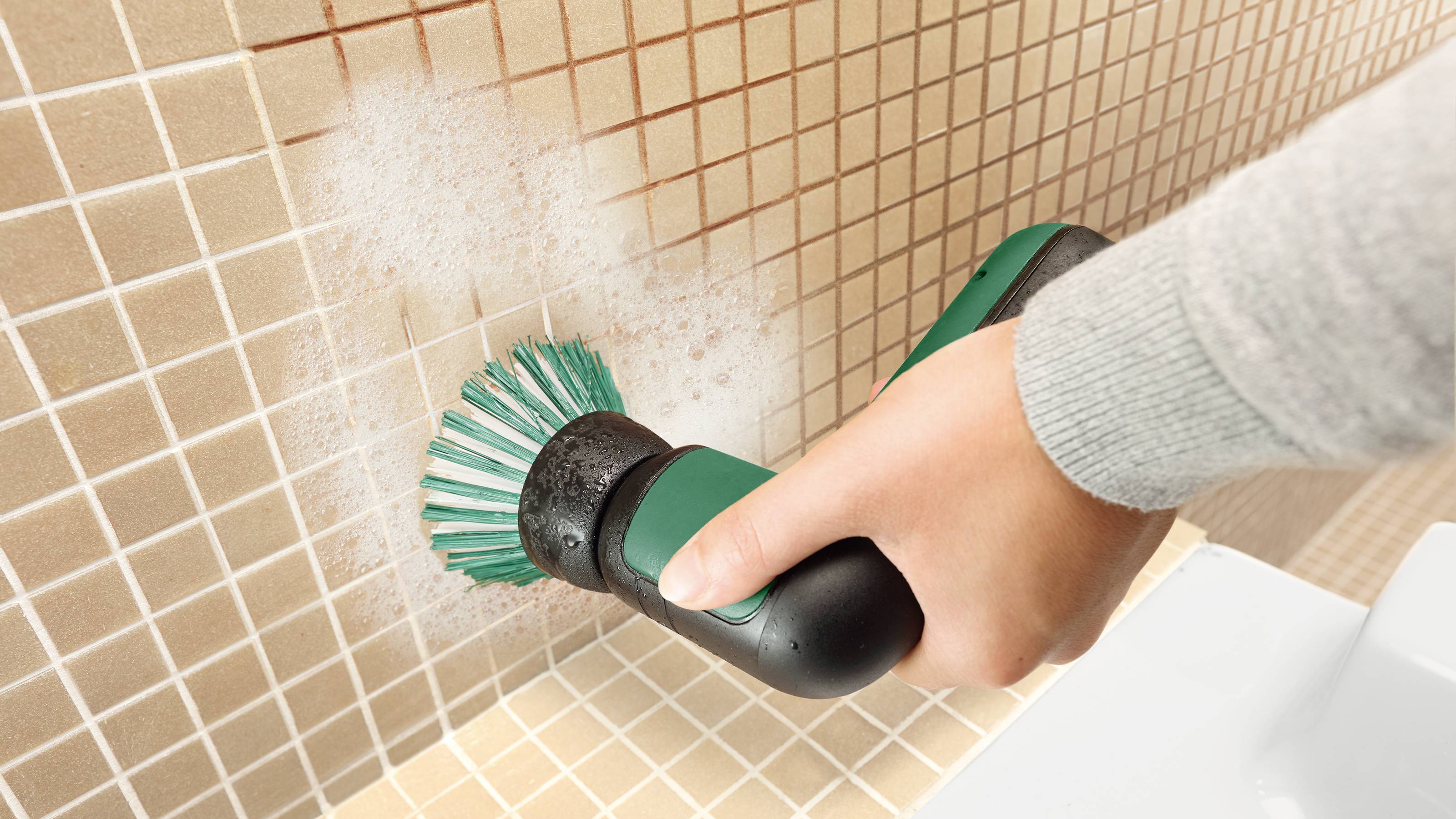 A person is cleaning wall-mounted tiles with an electric brush, generating soap foam.