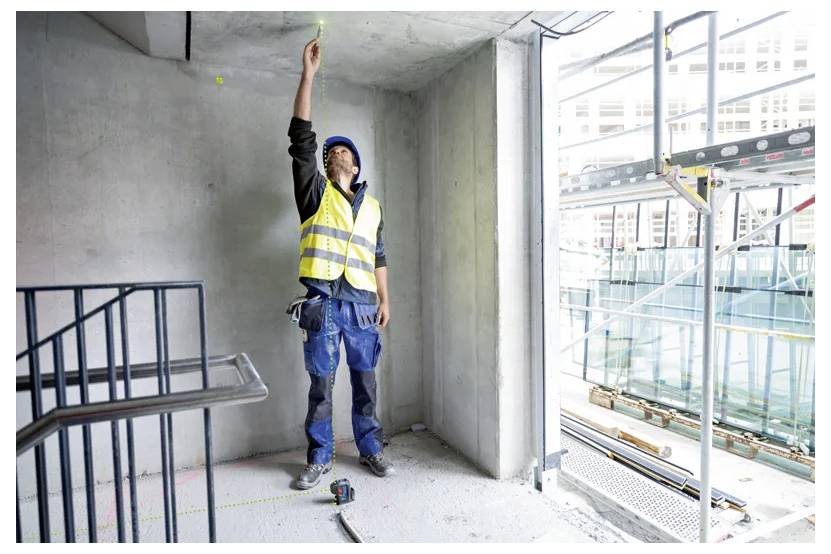 'A construction worker in a safety vest and helmet marks a ceiling point inside a partially constructed building with tools on the floor.'