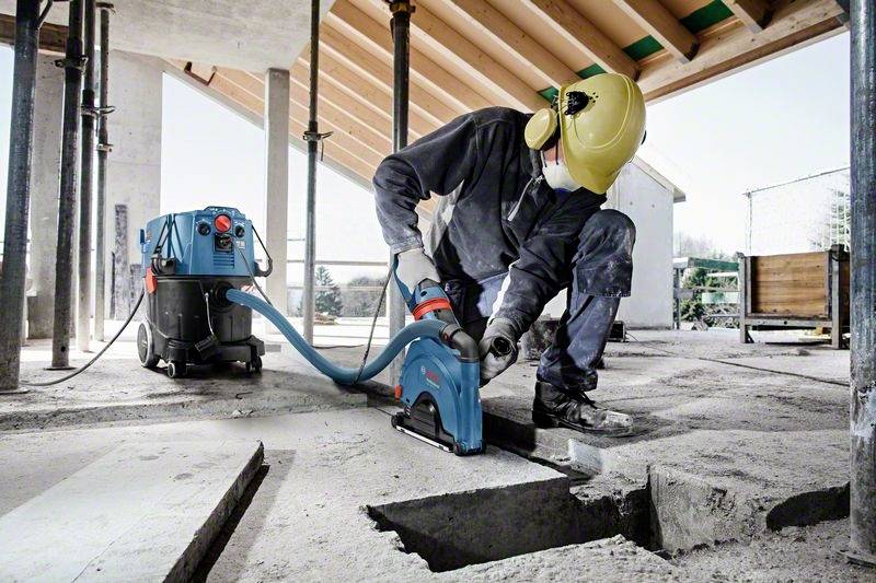 Construction worker wearing protective gear and using power tools to cut concrete on a building site. Modern building in the background.
