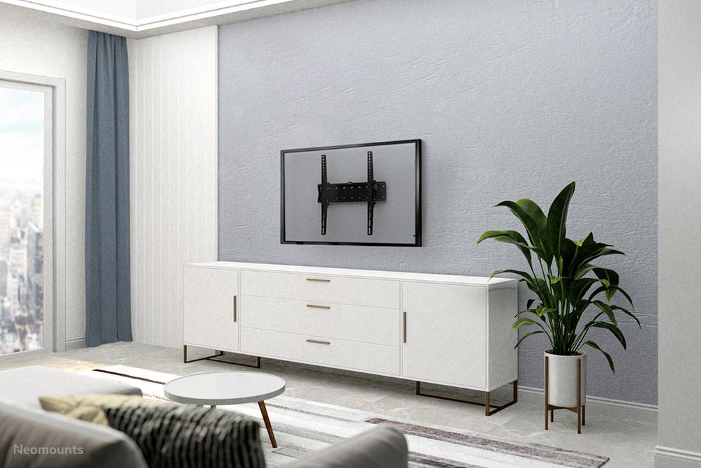 Simply furnished living room with a white sideboard, wall-mounted television without a screen, and a green plant. A city view can be seen through the window in the background.