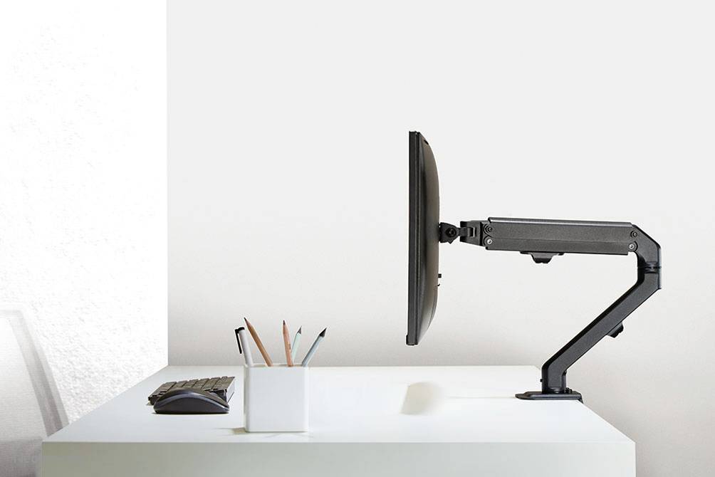 A monitor arm holds a screen to the side of a white desk, with a pen holder containing coloured pens beside it.