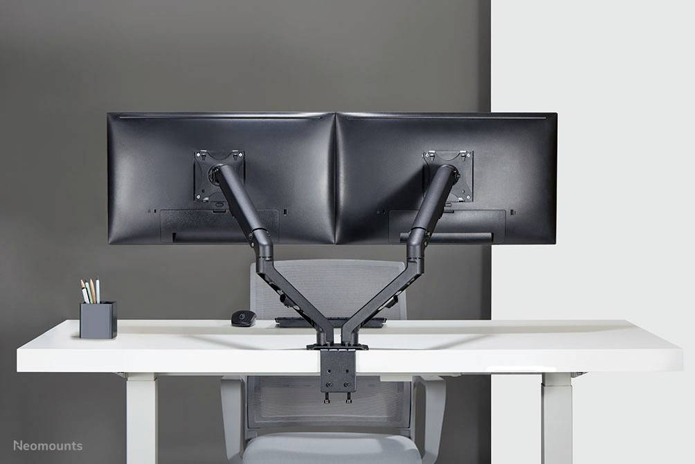 Two large monitors mounted on a double monitor arm on a desk. A grey wall is in the background.