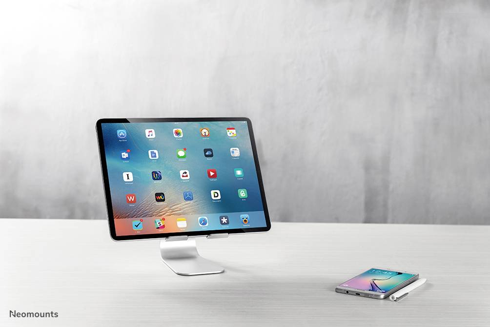 Tablet on a stand next to a smartphone on a table. Screen displays various app icons. Modern, minimalist look.