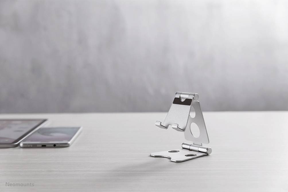 A silver, adjustable mobile phone holder stands on a desk; two smartphones lie in the background.