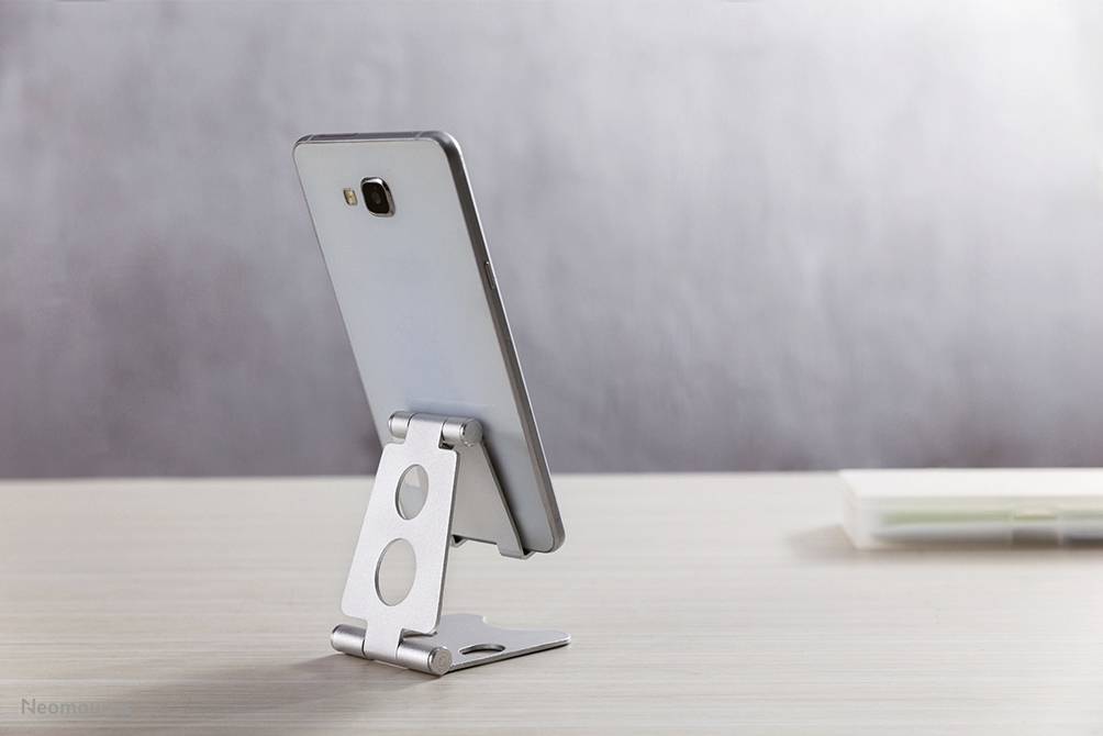 A smartphone on a table in a silver stand, viewed from the side. The background is blurred.