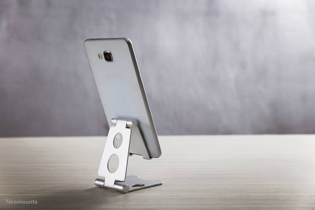 A smartphone sits in a silver, adjustable stand on a wooden table against a grey background.