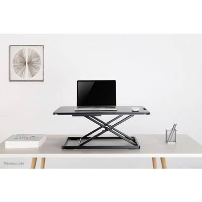 A height-adjustable desk riser with a laptop and a mouse placed on top. A piece of artwork and a pen holder are visible in the background.