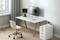 A modern workspace with a white desk, a monitor stand with screen, a keyboard, and a white chair on a wooden floor.