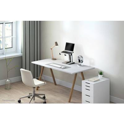 Modern desk with laptop stand, lamp, and headphones. White drawer beside it, bright environment with window and plants.