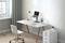 Modern desk with laptop stand, lamp, and headphones. White drawer beside it, bright environment with window and plants.